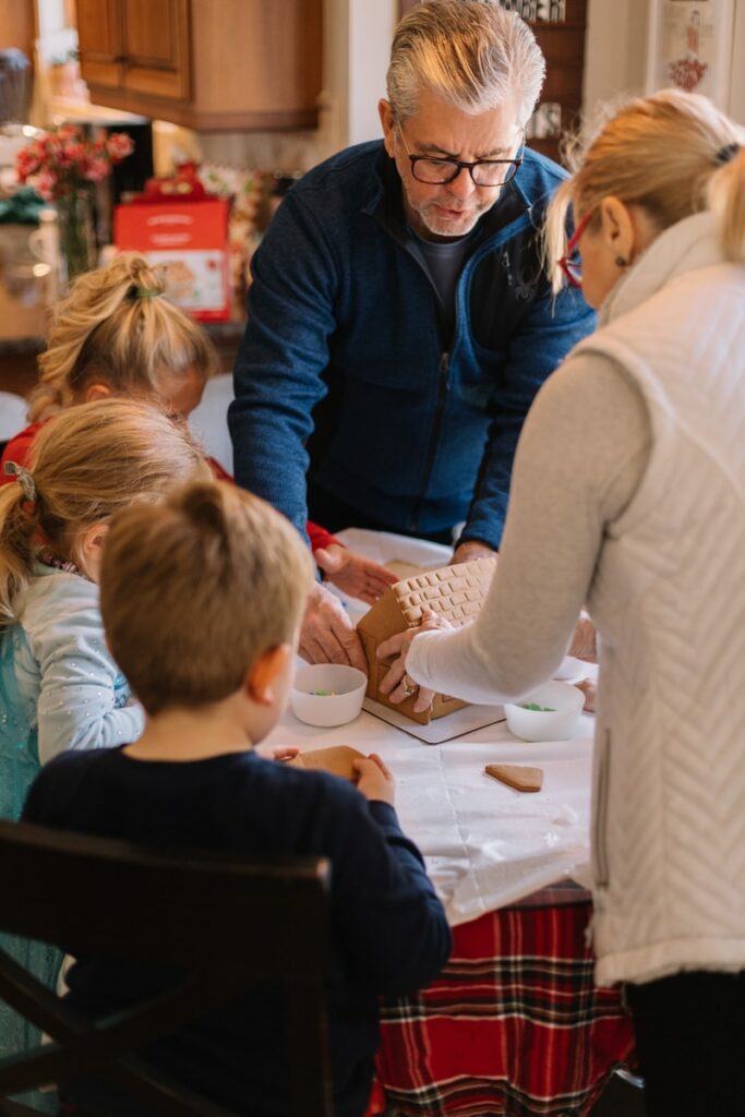 two women standing and three children sitting while preparing gingerbread house